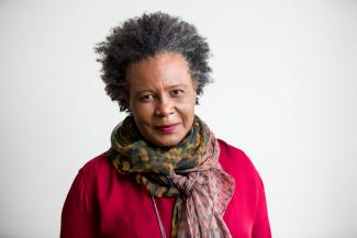 Claudia Rankine stands wearing a long-sleeved, fuchsia top and multicolored scarf against a blank wall