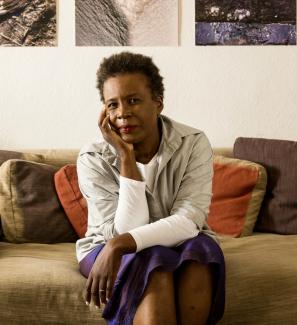 Claudia Rankine wears a long-sleeved beige top and iridescent purple, mid-length skirt. She sitss on a goldenrod sofa with rust and chocolate colored throw pillows. She rests her chin on her right hand, with her elbow against her knee.