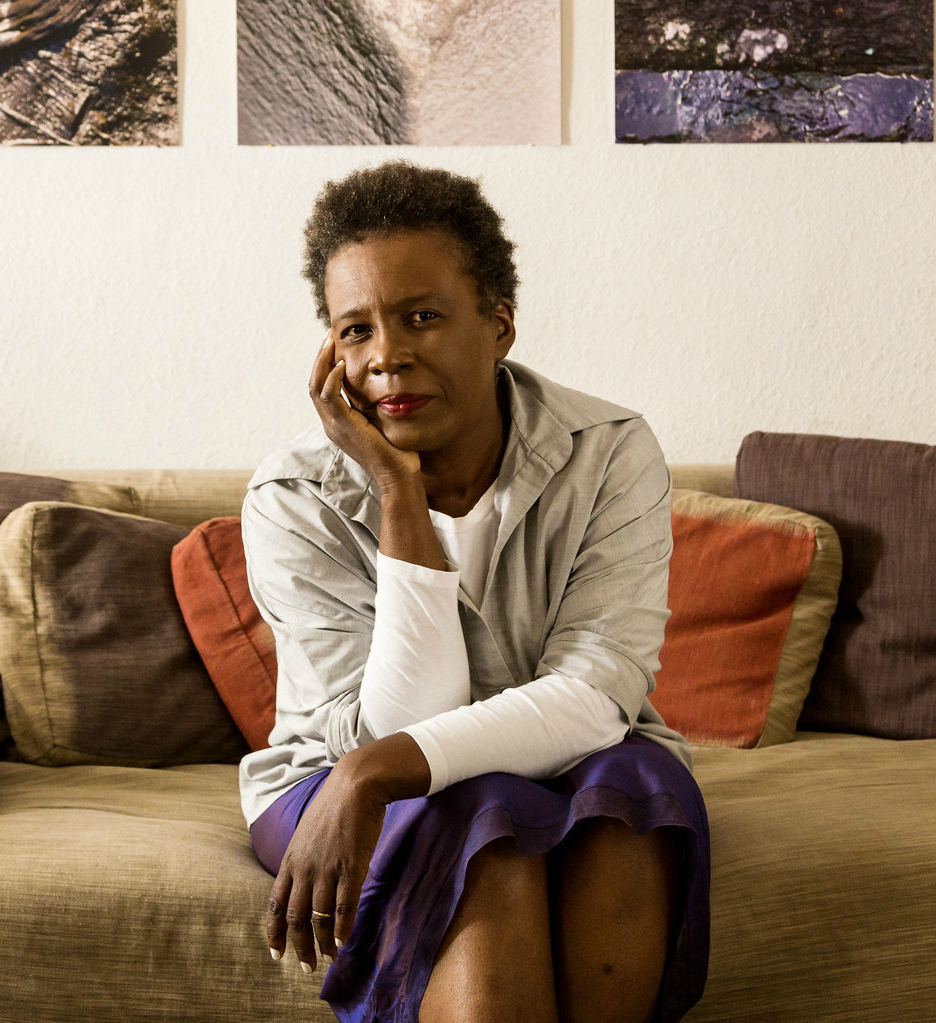 Claudia Rankine wears a long-sleeved beige top and iridescent purple, mid-length skirt. She sitss on a goldenrod sofa with rust and chocolate colored throw pillows. She rests her chin on her right hand, with her elbow against her knee.